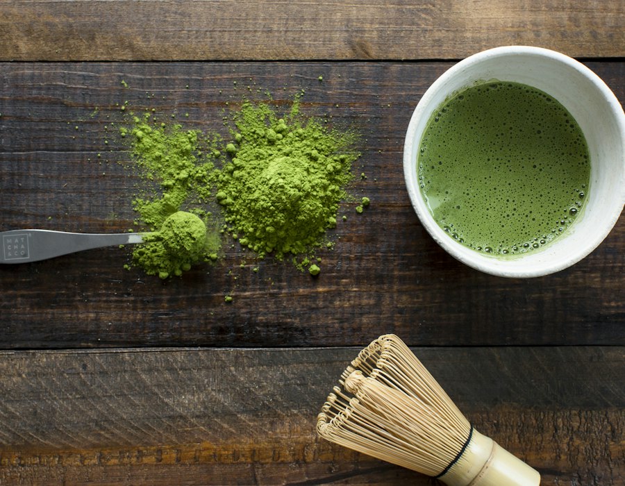 Bright green matcha powder next to a bowl of whisked matcha and a bamboo whisk
